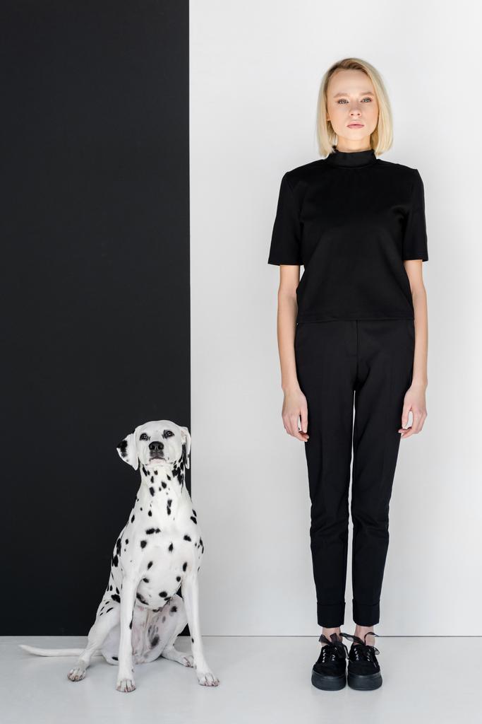 houkutteleva tyylikäs blondi nainen mustissa vaatteissa seisoo lähellä mustavalkoista seinää dalmatialaisella koiralla
 - Valokuva, kuva