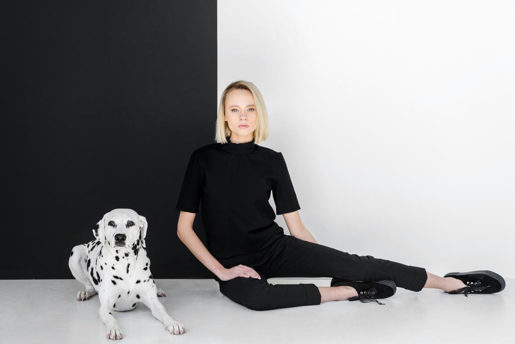 atraktivní stylové blond žena v černých šatech sedí poblíž černé a bílé zdi se Dalmatin pes - Fotografie, Obrázek