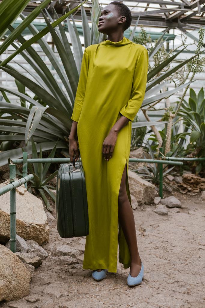 stylische Afroamerikanerin in gelbem Kleid posiert mit Reisetasche im tropischen Gewächshaus - Foto, Bild