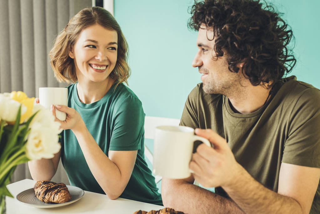 happy couple holding cups and smiling each other while having breakfast together - Photo, Image