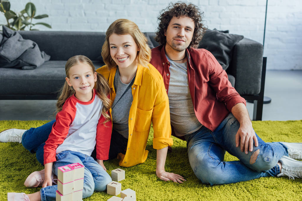 gelukkige familie zittend op een tapijt en glimlachend in de camera tijdens het spelen met houten blokken thuis - Foto, afbeelding