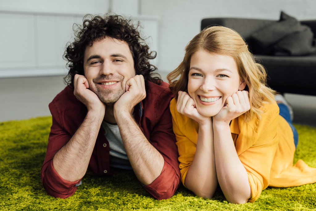 hermosa feliz pareja acostada en la alfombra y sonriendo a la cámara en casa
 - Foto, Imagen
