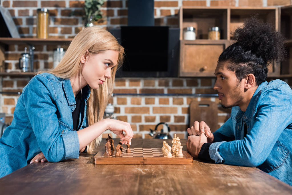 вид сбоку на многонациональную молодую пару, играющую вместе в шахматы на кухне
 - Фото, изображение