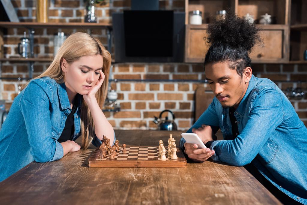 puolella näkymä kyllästynyt valkoihoinen nainen ja afrikkalainen amerikkalainen mies älypuhelin pöydässä shakki keittiössä
 - Valokuva, kuva