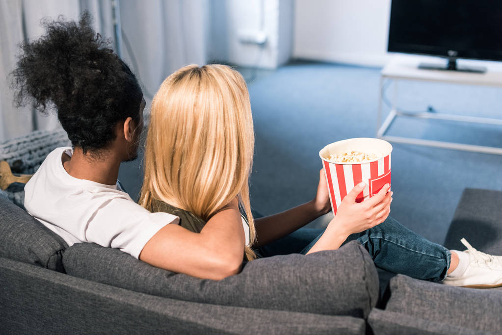 back view of multiracial couple with popcorn watching film together at home - Photo, Image
