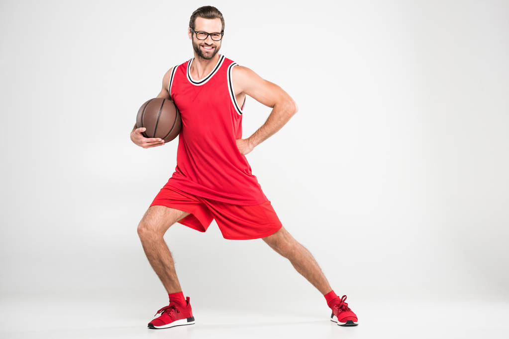 Lächelnder Basketballspieler in roter Sportbekleidung und Retro-Brille posiert mit Ball, isoliert auf weiß - Foto, Bild