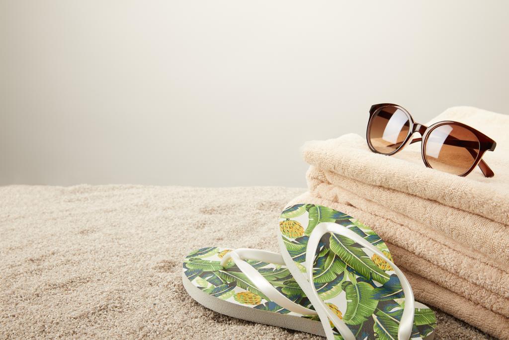 vista de cerca de la pila de toallas, gafas de sol y chanclas de verano sobre arena sobre fondo gris
 - Foto, Imagen
