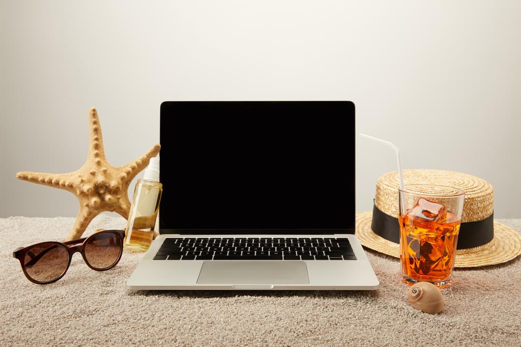 Nahaufnahme von Laptop mit leerem Bildschirm, erfrischendem Cocktail und Sommerurlaub-Accessoires auf Sand auf grauem Hintergrund - Foto, Bild