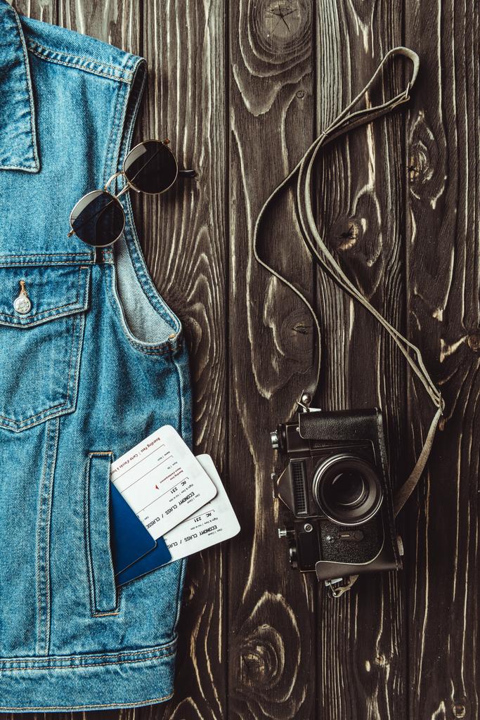 квартира с джинсовой жилеткой, паспортами, билетами, солнцезащитными очками и ретро фотокамерой на темной деревянной поверхности
 - Фото, изображение