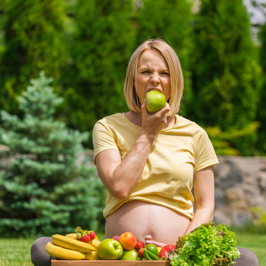Mulher grávida sentada na grama e segurando uma maçã na mão
 - Foto, Imagem