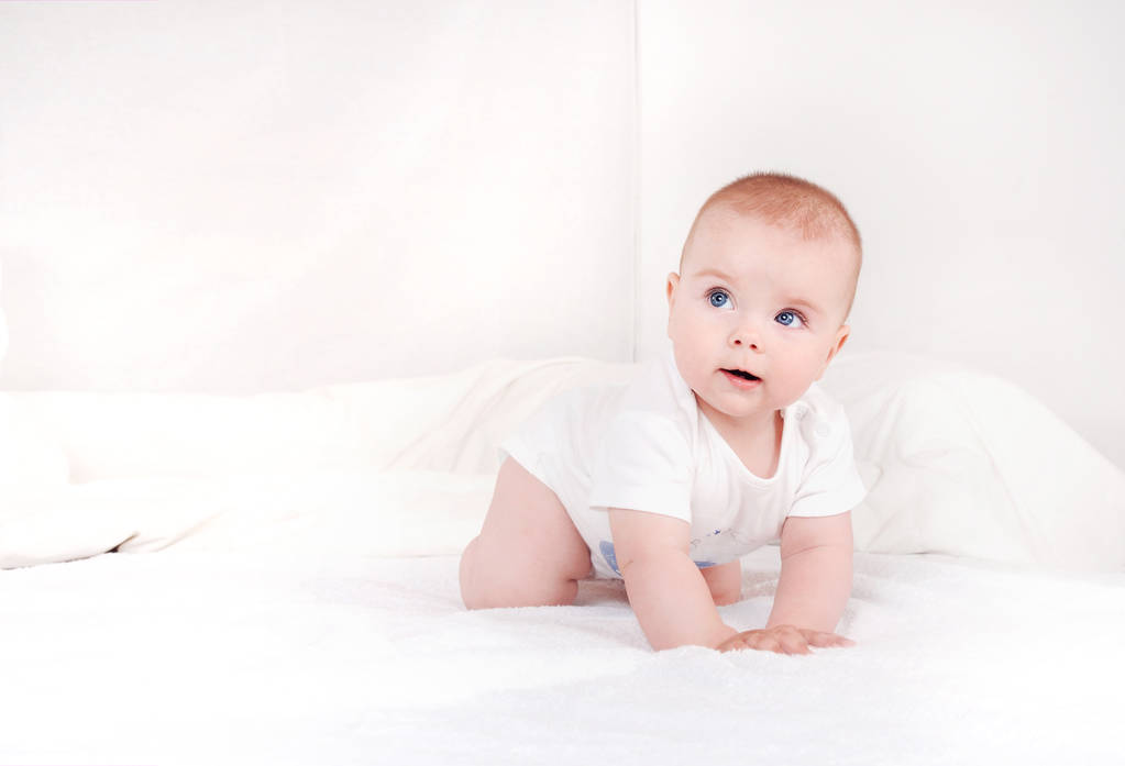 Portré a édes újszülött csecsemő crawlng a fehér ágy súlyos keres el. Boldog mosolygó gyermek szeme kék háttér fehér ágy - Fotó, kép