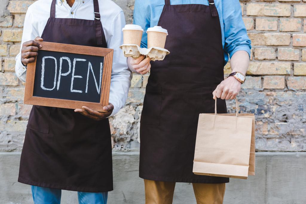 abgeschnittene Aufnahme zweier multiethnischer Coffeeshop-Besitzer mit offenem Schild, Papiertüten und Einweg-Kaffeetassen, während sie in der Nähe einer Mauer stehen - Foto, Bild