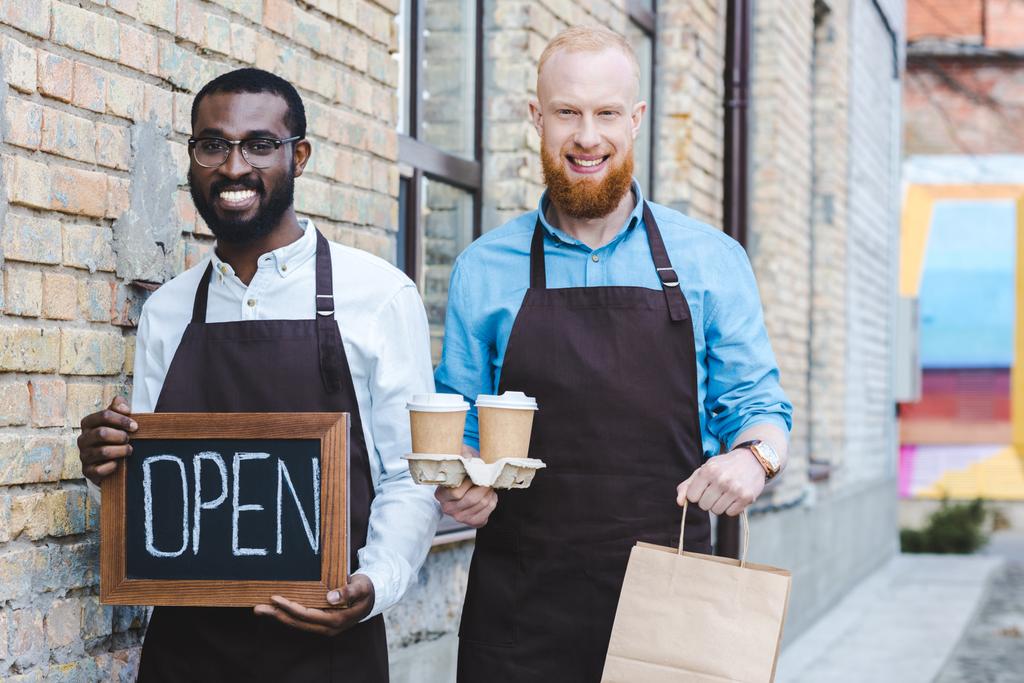 junge gutaussehende multiethnische Coffeeshop-Besitzer in Schürzen mit offenen Schildern, Papiertüten und Einweg-Kaffeetassen lächeln in die Kamera  - Foto, Bild