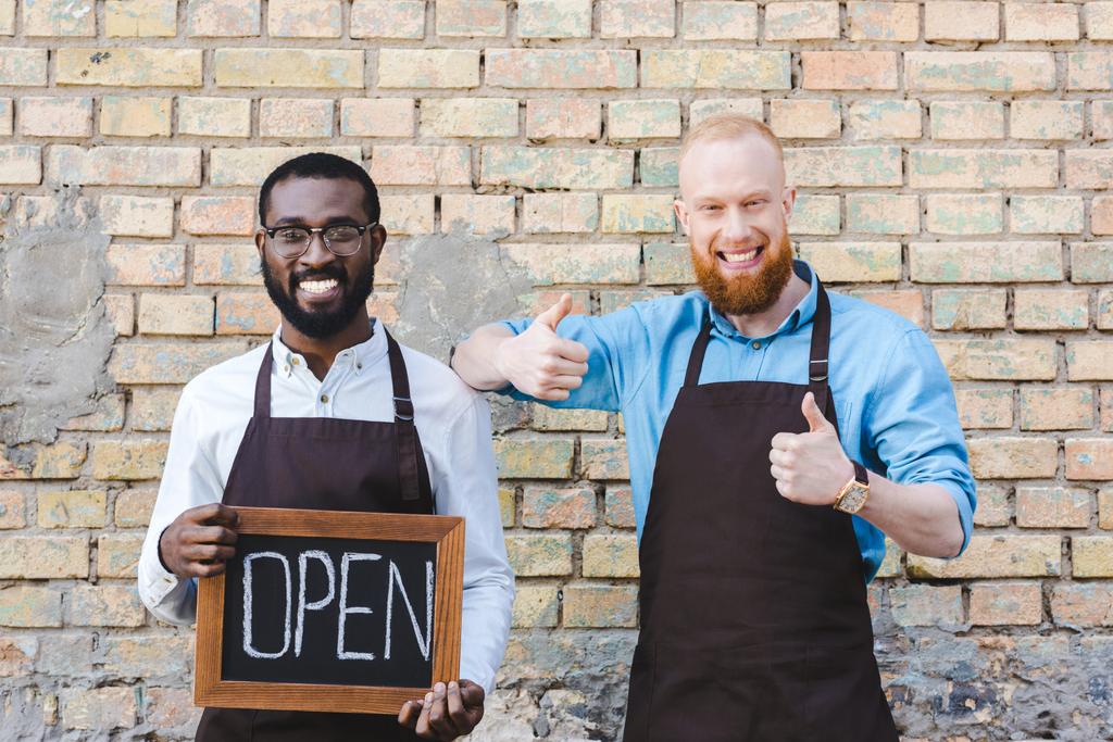 handsome multiethnic owners of coffee shop in aprons holding sign open and showing thumbs up, smiling at camera - Photo, Image