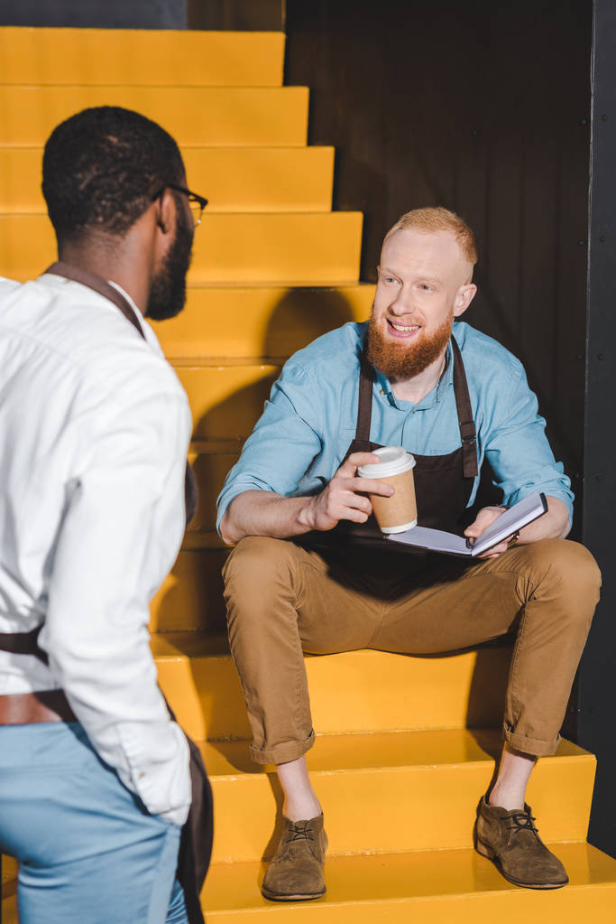 two multiethnic male baristas looking at each other and discussing work on stairs - Photo, Image