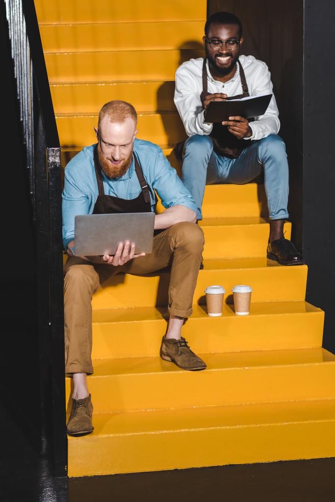 zwei junge lächelnde Coffeeshop-Besitzer in Schürzen mit Laptop und Lehrbuch sitzen auf der Treppe mit Pappbechern Kaffee  - Foto, Bild