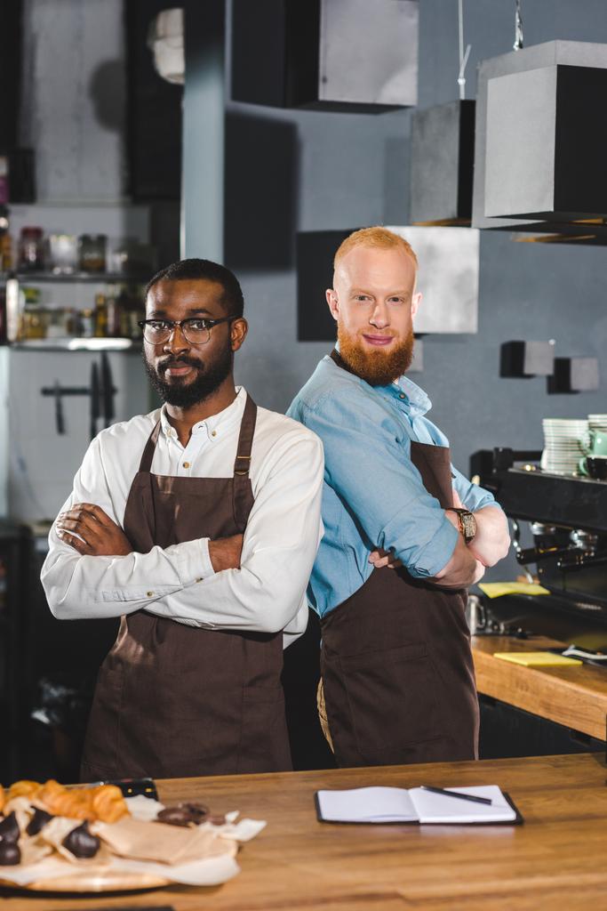 два молодых мультикультурных бармена в фартуках, стоящих со скрещенными руками в кофейне
 - Фото, изображение
