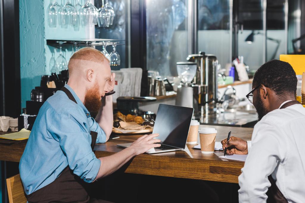 visão traseira de jovens proprietários multiétnicos do sexo masculino de café que trabalham à mesa com livro didático e laptop
  - Foto, Imagem