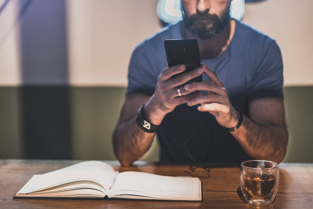 Молодий бородатий чоловік сидить у кафе і використовує мобільний телефон для прослуховування цифрової музики. Чоловік робить нотатки в записнику. Горизонтально. Розмитий фон
. - Фото, зображення