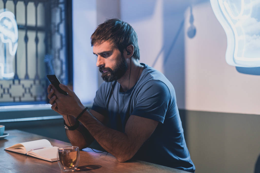 Молодой бородатый мужчина сидит в кафе и использует смартфон для прослушивания аудиокниги. Человек делает заметки на блокноте. Горизонтально. Размытый фон
. - Фото, изображение