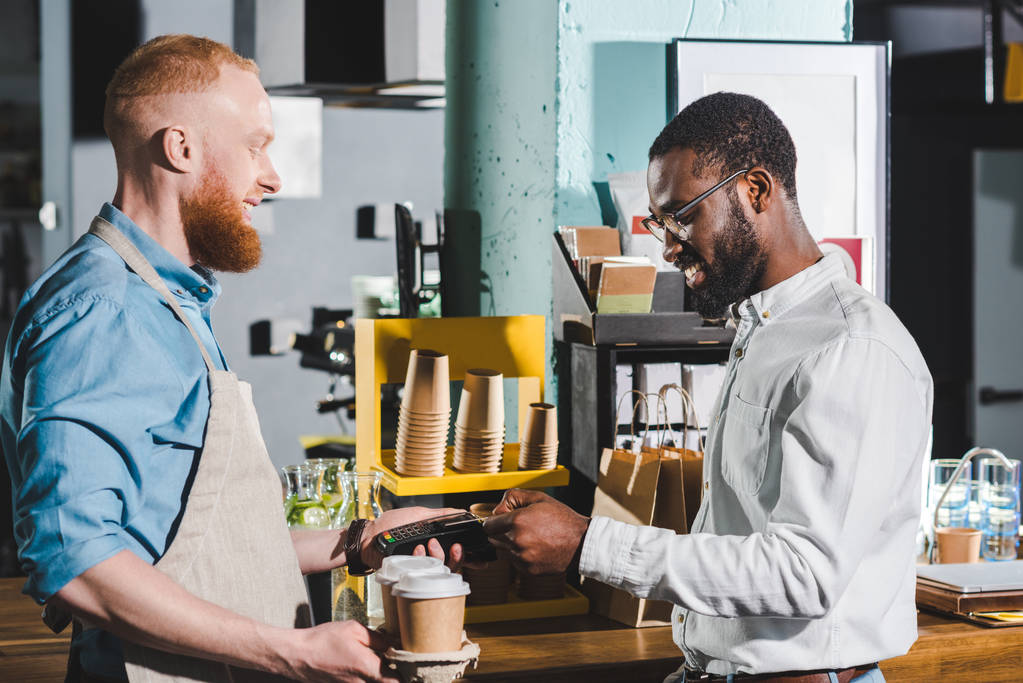 Африканский американец платит кредитной картой и улыбается мужчина-бариста с терминалом и бумажными чашками кофе
 - Фото, изображение