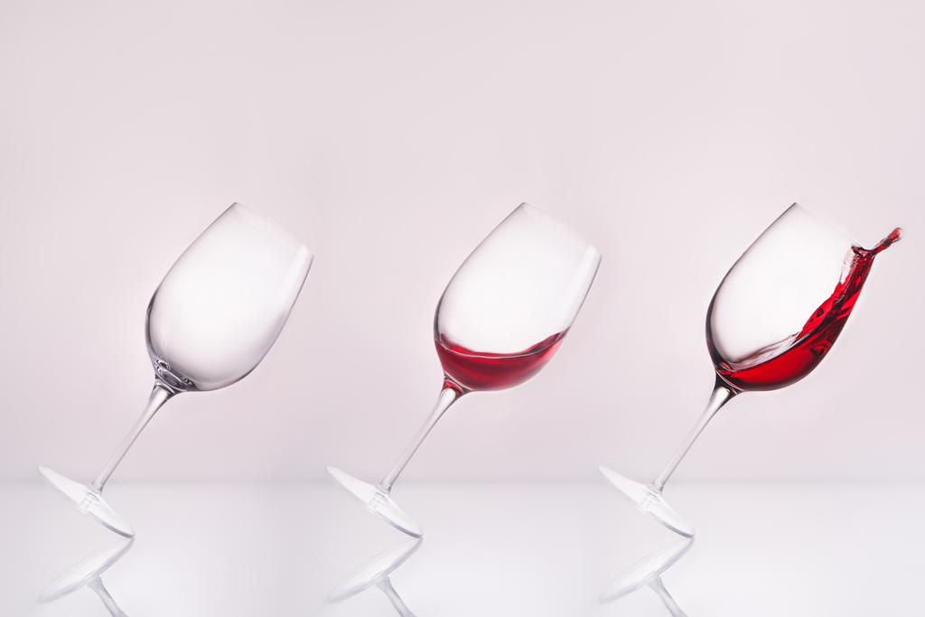 row of inclined wineglasses empty and with splashing wine on reflective surface and on white - Photo, Image