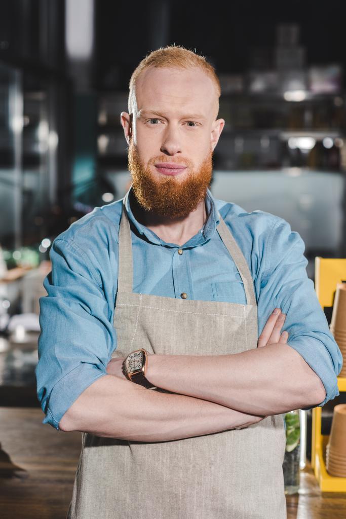 jonge mannelijke barista in schort staande met gekruiste armen in coffeeshop - Foto, afbeelding
