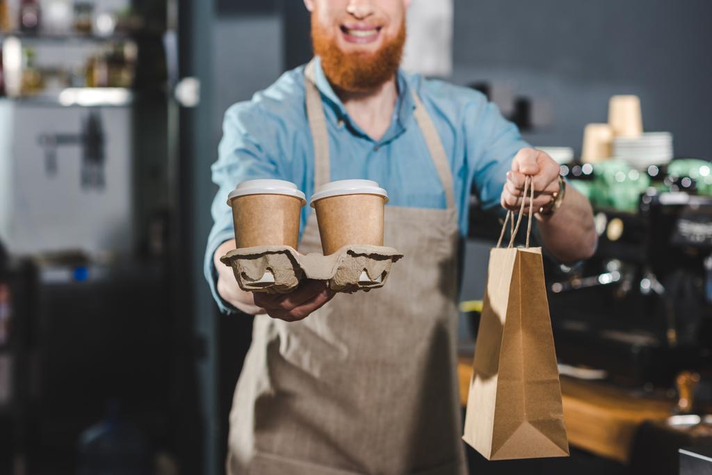 bijgesneden schot van lachende bebaarde barista holding wegwerp koffie cups en papieren zak in café - Foto, afbeelding