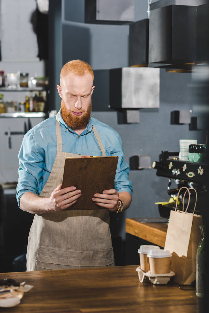serious bearded barista in apron holding clipboard while working in coffee shop - Photo, Image