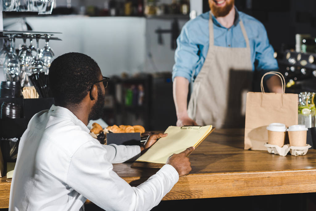 african american client holding blank clipboard and looking at bearded barista in coffee shop - Photo, Image