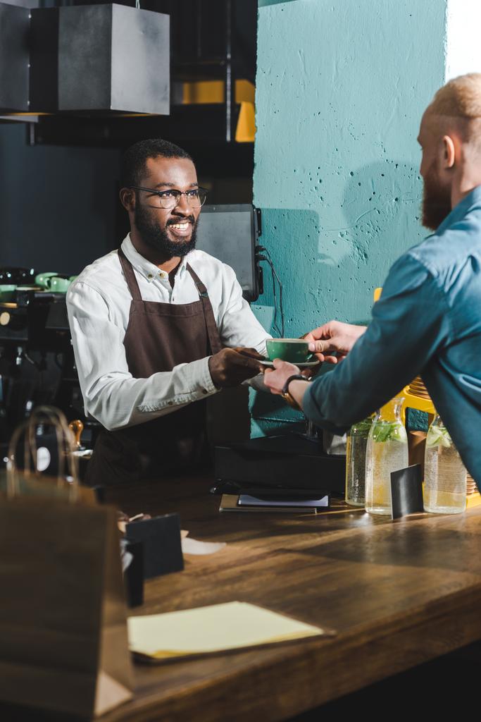 cropped shot of cheerful african american barista giving cup of coffee to bearded young client in cafe - Photo, Image