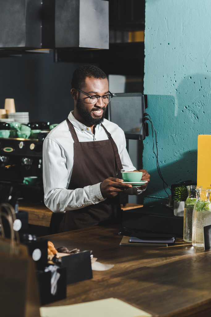 カフェでのコーヒーのカップを保持している眼鏡でハンサムな笑顔のアフリカ系アメリカ人バリスタ - 写真・画像