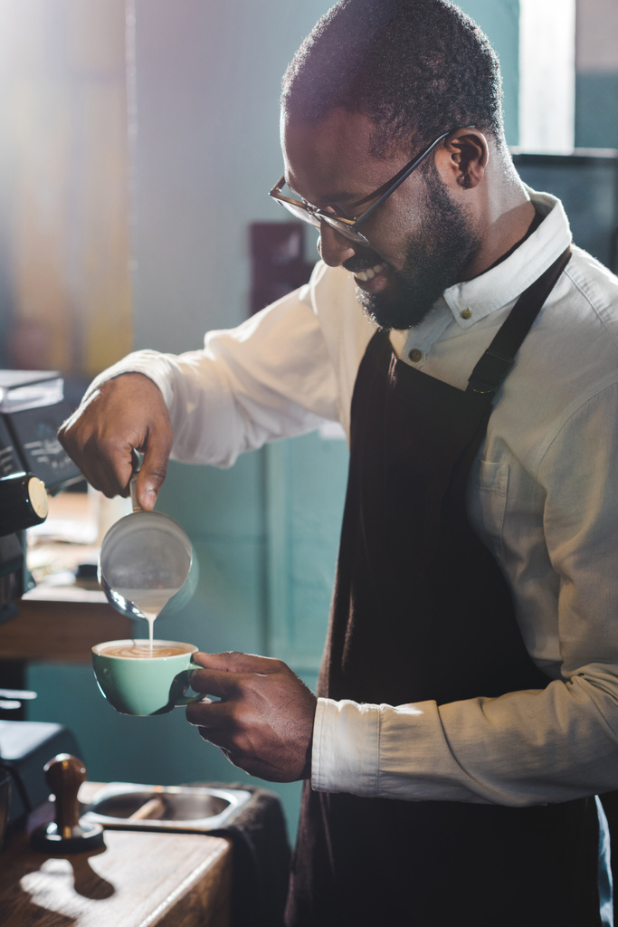 smiling young african american barista in eyeglasses pouring milk while making cappuccino - Photo, Image