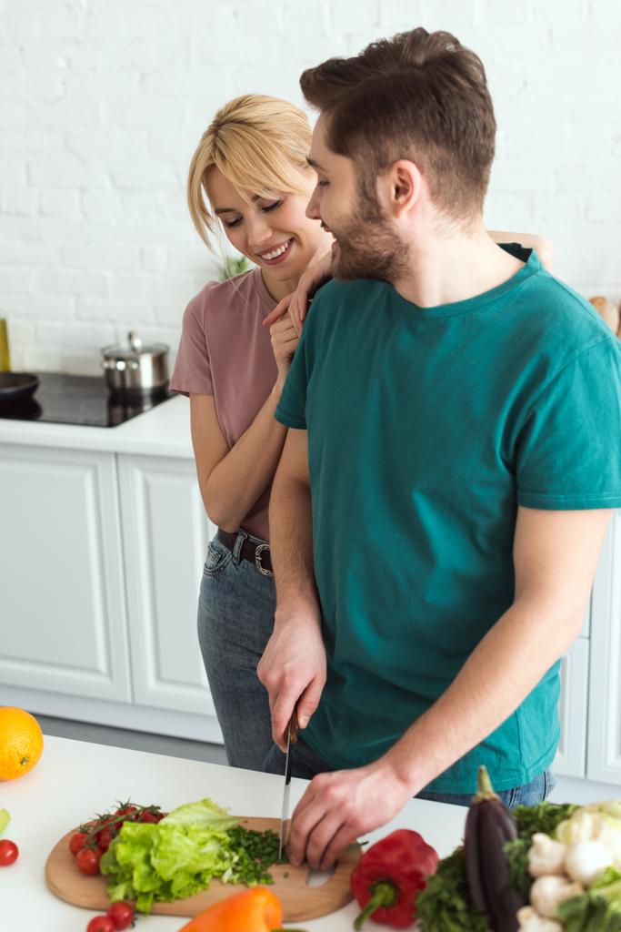 vegan boyfriend cutting vegetables and girlfriend hugging him at kitchen - Photo, Image