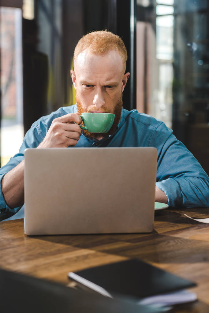 Knappe jongeman die koffie drinkt en laptop gebruikt in café - Foto, afbeelding