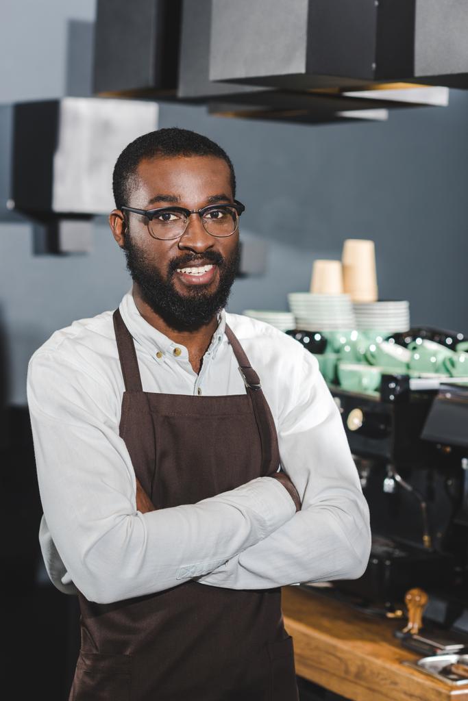 αυτοπεποίθηση αφρικανική αμερικανική barista σε γυαλιά στέκεται με σταυρωμένα χέρια και να χαμογελάει κάμερα σε καφενείο - Φωτογραφία, εικόνα