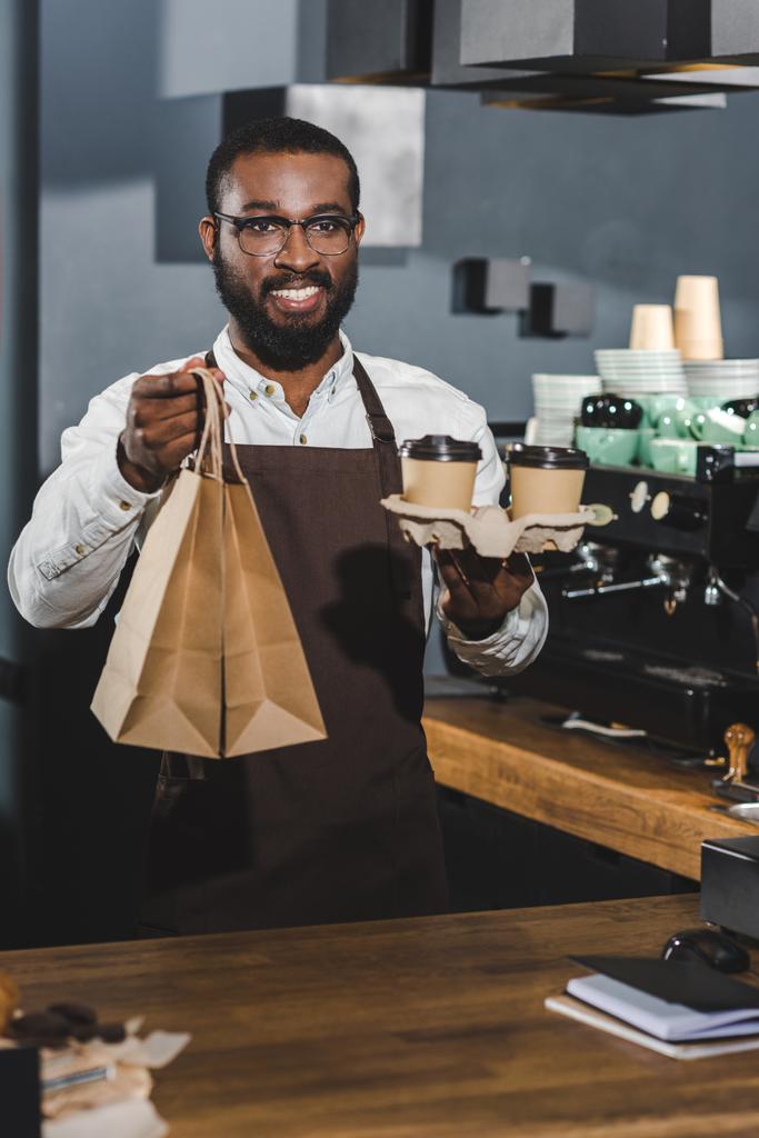 beau sourire jeune afro-américain barista tenant tasses à café jetables et sacs en papier dans un café
  - Photo, image