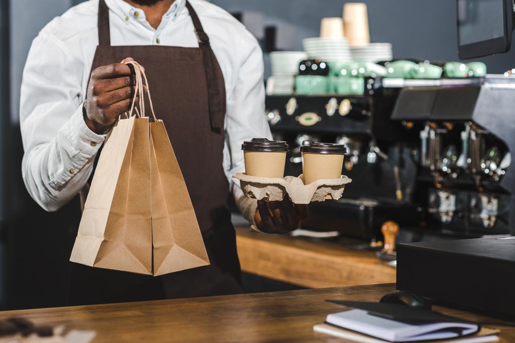 foto recortada de barista afroamericano sosteniendo tazas de café desechables y bolsas de papel en la cafetería
 - Foto, Imagen