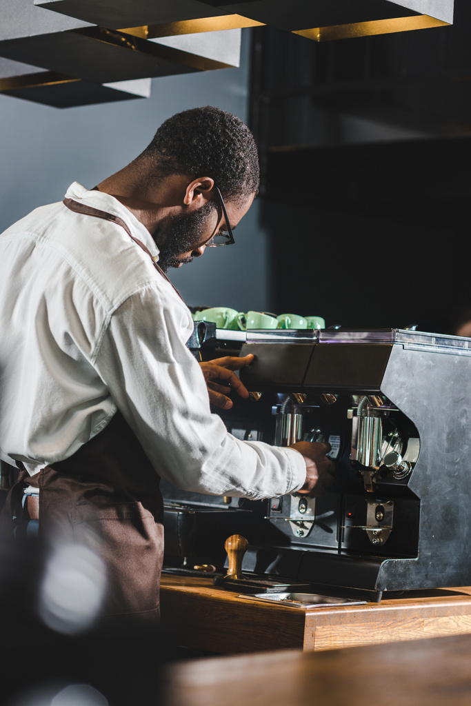 眼鏡とコーヒー マシンの操作のエプロンの若いアフリカ系アメリカ人バリスタ - 写真・画像