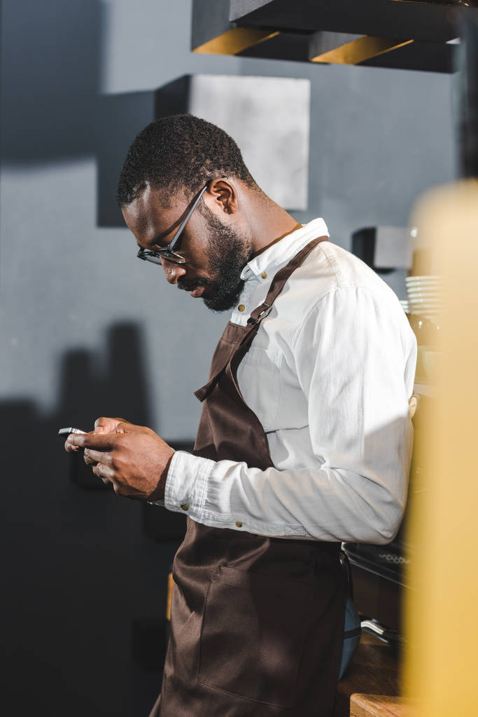 side view of young african american bartender using smartphone in coffee shop - Photo, Image