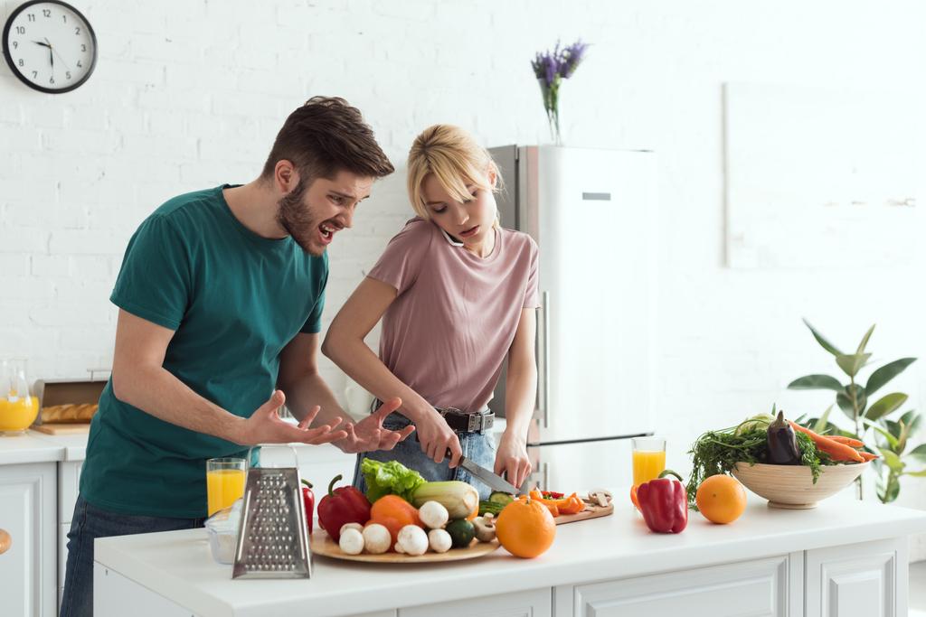 přítel, šklebící se zároveň přítelkyně vaření a mluví o smartphone v kuchyni - Fotografie, Obrázek