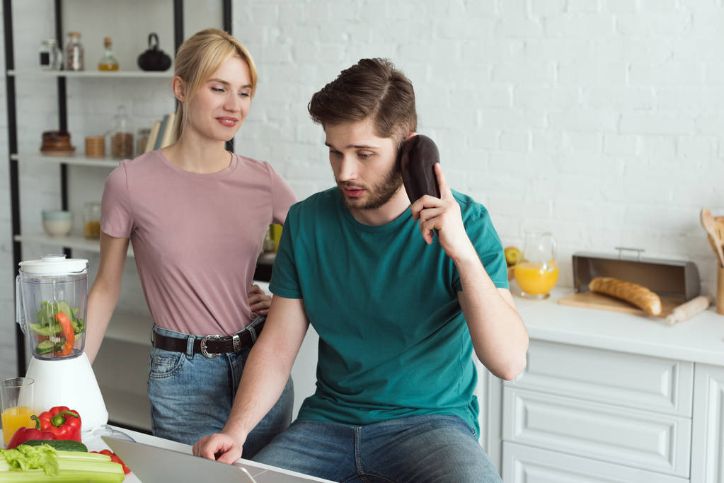 man pretending talking on eggplant at table with laptop and girlfriend near by in kitchen at home, vegan lifestyle concept - Photo, Image