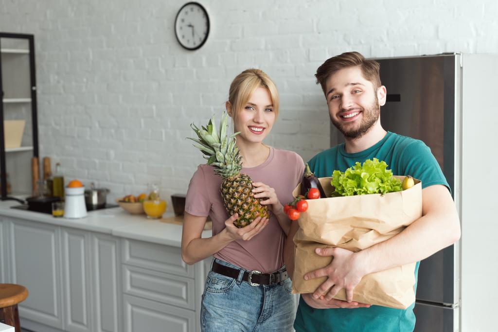 couple végétalien souriant avec sac en papier plein de légumes frais dans la cuisine à la maison
 - Photo, image