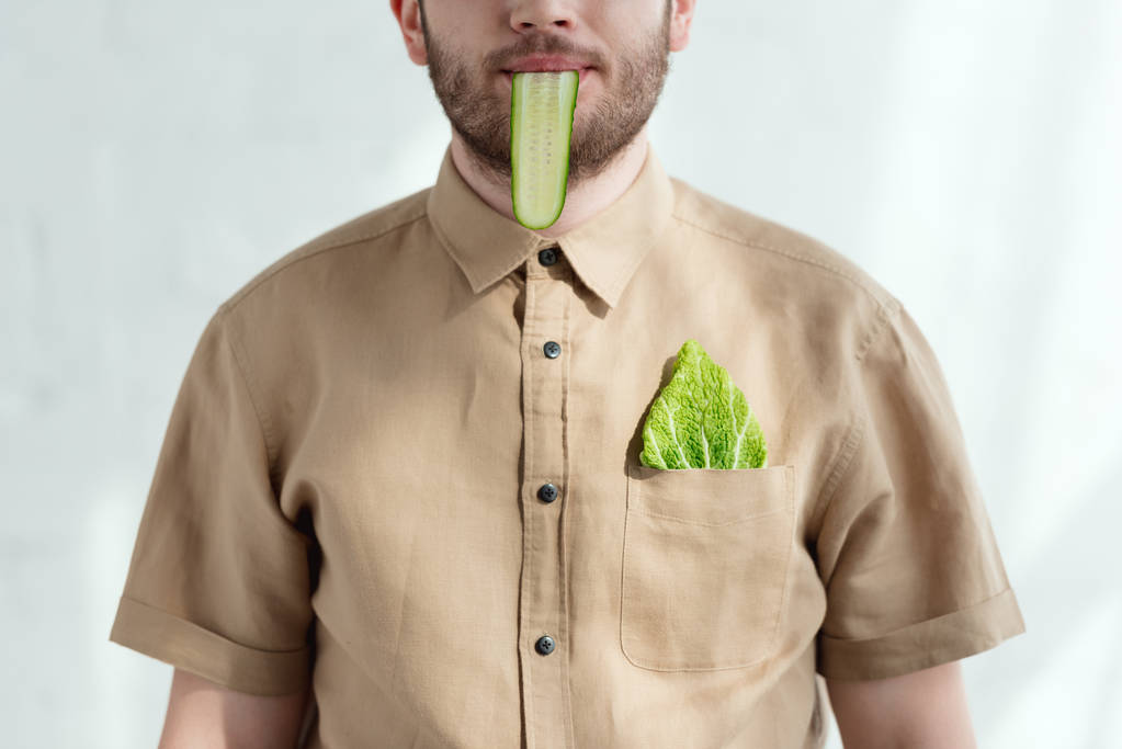 Μερική άποψη του ανθρώπου με φέτα αγγούρι στο στόμα και savoy λάχανο φύλλο στην τσέπη, έννοια τρόπου ζωής για χορτοφάγους  - Φωτογραφία, εικόνα