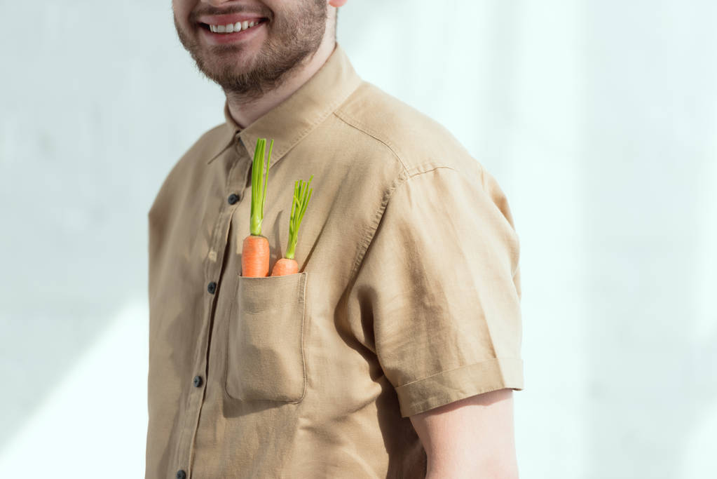 bijgesneden schot van lachende bebaarde man met verse wortelen in zak, veganistische levensstijl concept - Foto, afbeelding