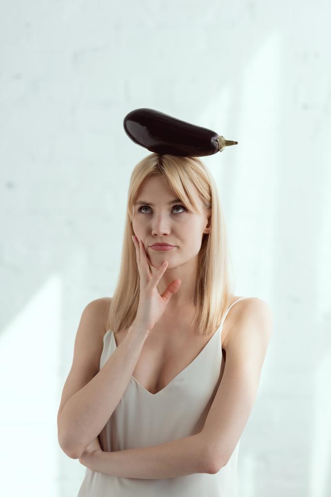 Portret van peinzende jongedame met verse aubergine op hoofd, veganistische levensstijl concept - Foto, afbeelding