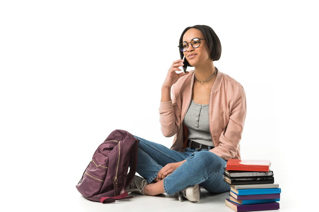 estudiante afroamericana hablando por teléfono inteligente y sentada con mochila y libros en el suelo, aislada en blanco
 - Foto, imagen