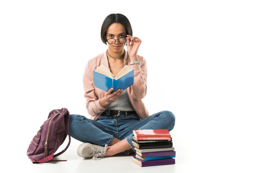 feminino afro-americano estudante em óculos lendo livros enquanto sentado no chão com mochila, isolado no branco
  - Foto, Imagem