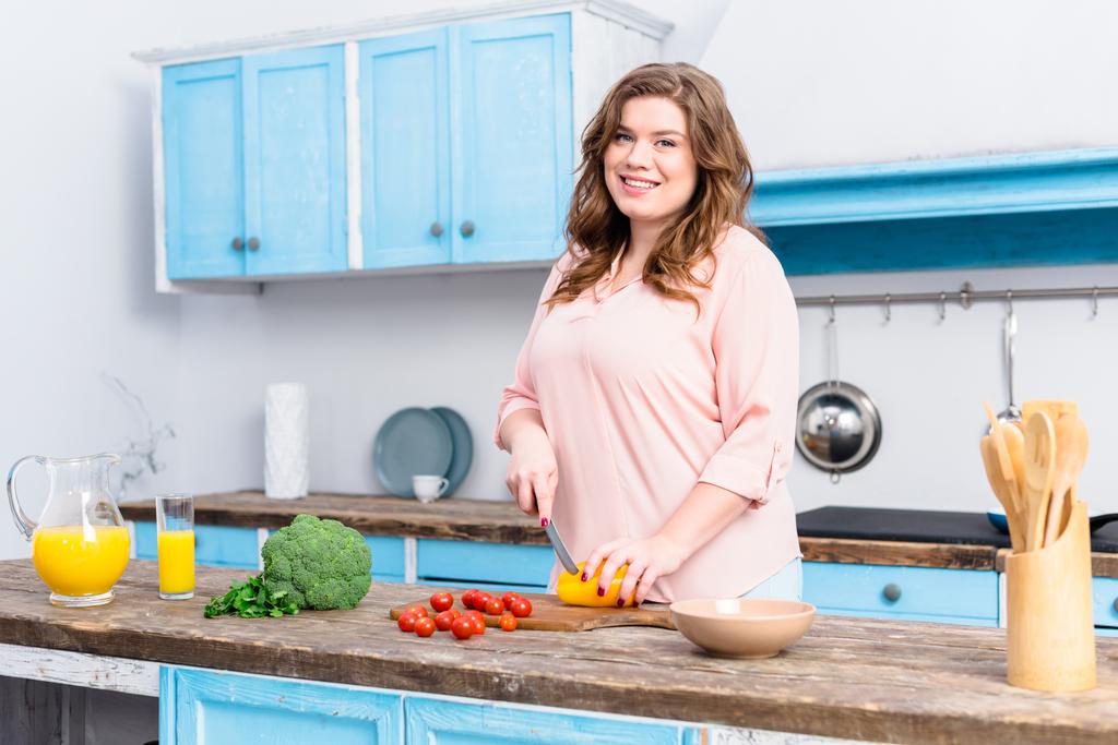 Übergewichtige junge lächelnde Frau schneidet Gemüse für Salat in der heimischen Küche  - Foto, Bild