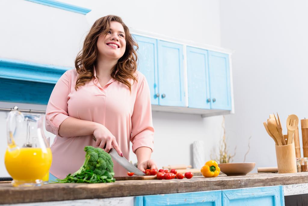 ylipainoinen nuori hymyilevä nainen leikkaa vihanneksia salaattia keittiössä kotona
 - Valokuva, kuva
